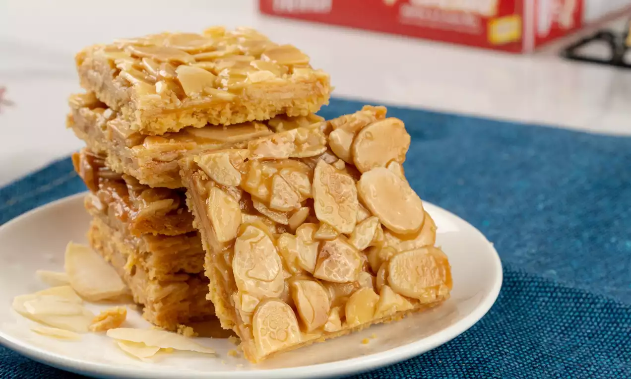 Stack of golden-brown salted caramel almond pancake cookies topped with glossy almond slices on a white plate with scattered flakes