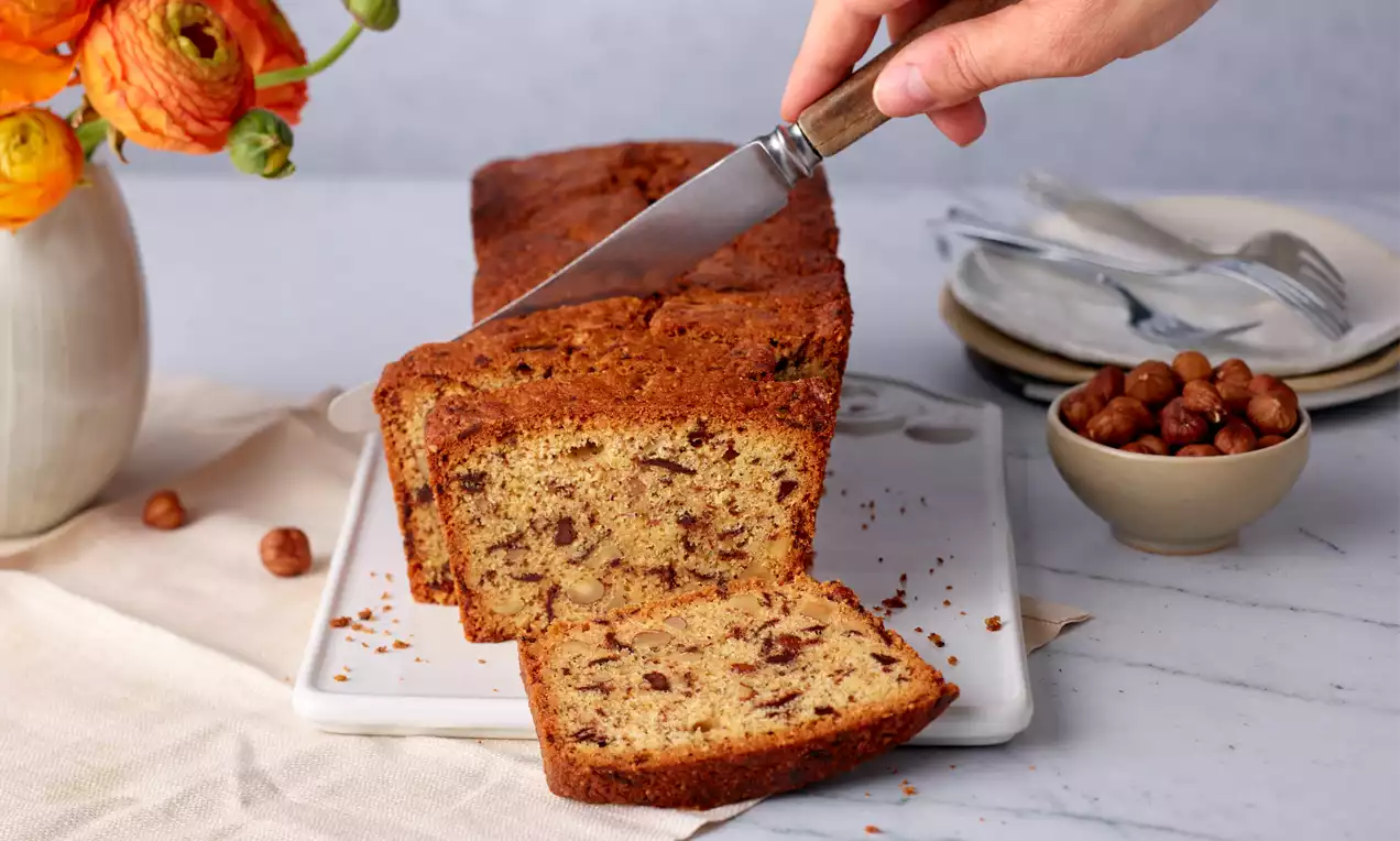 Saftiger Nusskuchen mit goldbrauner Kruste, in Scheiben geschnitten auf weißer Platte, umgeben von Haselnüssen und dekorativem Geschirr