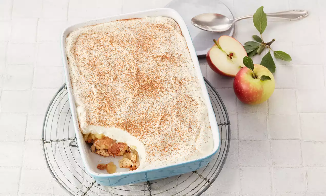 Apfel-Tiramisu in rechteckiger Form mit heller Mascarpone-Creme, bestäubt mit Zimt, daneben frische Äpfel und ein Löffel auf weißem Untergrund