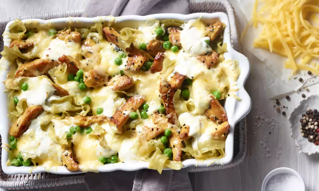 Goldbraune Hähnchenstücke, zart geschmolzener Käse, grüne Erbsen und helle Soße in einer weißen Auflaufform auf einem grauen Stoff