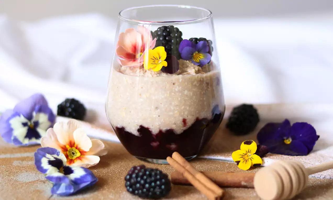 Ovesná kaše světle béžové barvy ve sklenici s vrstvou tmavě fialového pyré, ozdobená čerstvými ostružinami, jedlými květy a skořicovými tyčinkami