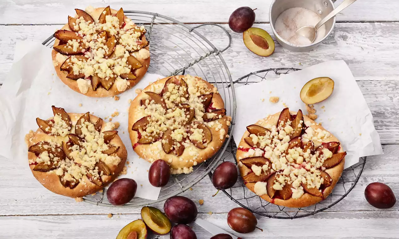 Okrúhle nadýchané koláče zo zlatistého cesta, zdobené plátkami tmavofialových sliviek a posypané svetlou mrvenicou na mriežke s čerstvými slivkami