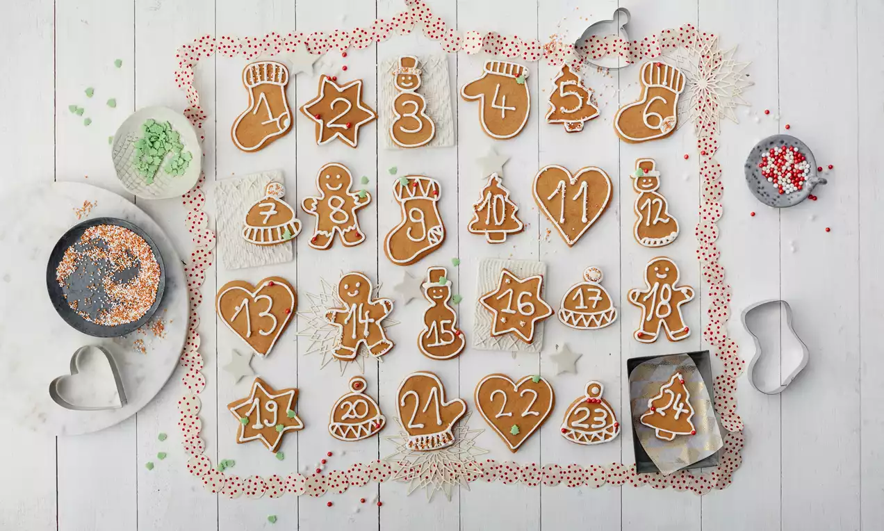 Adventskalender aus goldbraunen Plätzchen in verschiedenen Formen mit weißem Zuckerguss verziert auf hellem Holz mit Backdekoration