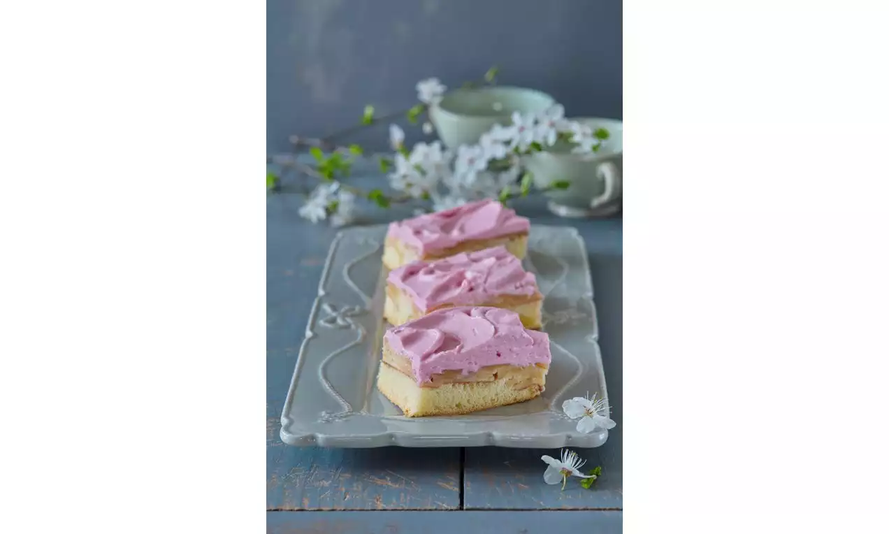 Tři obdélníkové řezy obráceného jablečníku na zdobeném podnosu, s vrstvou růžové pěny a světlého těsta, obklopené bílými kvítky