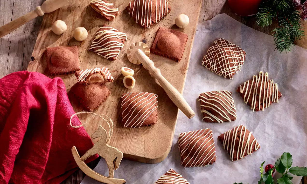 Ravioli-förmige braune Kekse mit weißer Zuckerglasur verziert, auf einem Holzbrett und Backpapier arrangiert, umgeben von roten und grünen Dekorationen