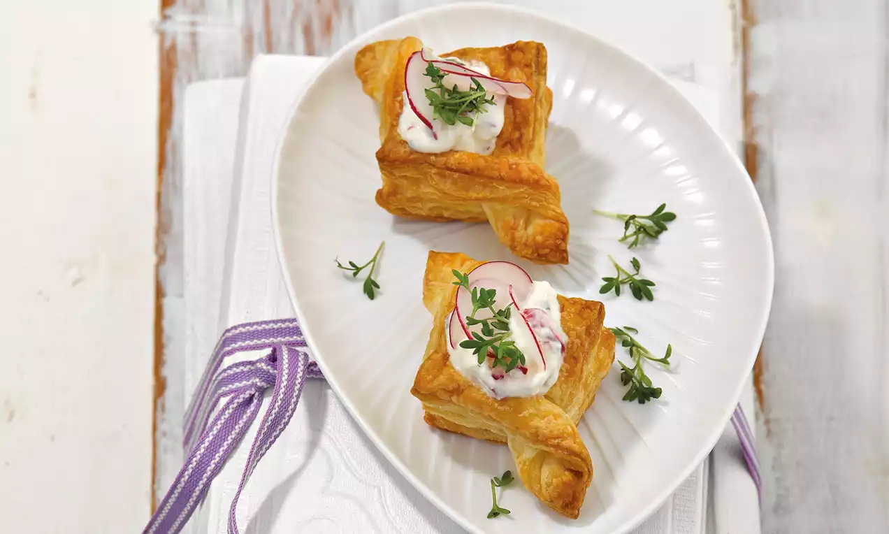 Aranybarna, ropogós leveles tészta négyzetek kettő krémes, fehér öntettel, retek szeletekkel és friss zölddel díszítve fehér tányéron