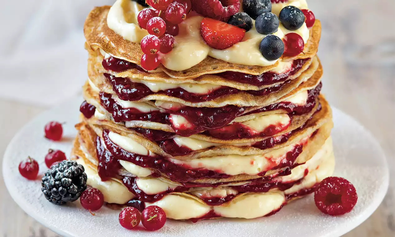 Vrstvený palacinkový tort s jemne zlatistými okrajmi, plnený vanilkovým krémom a ovocným džemom, ozdobený čerstvými plodmi na bielom tanieri