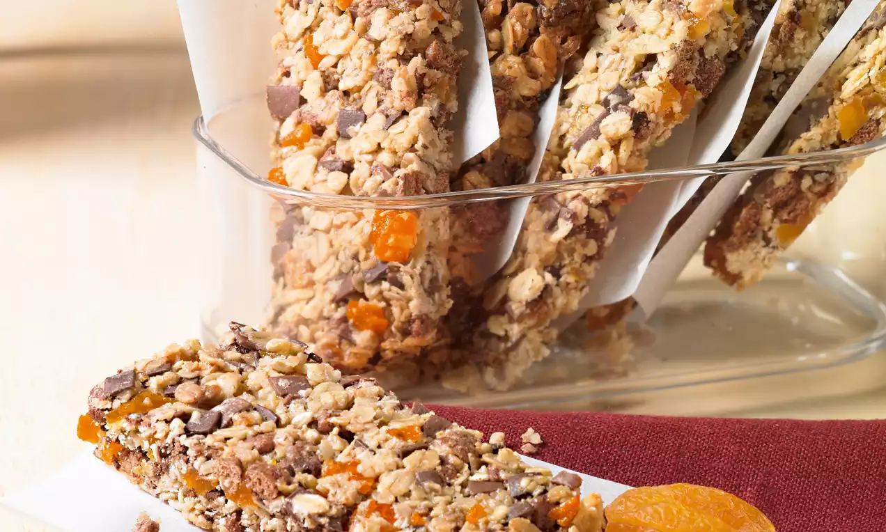 Vitalis marelične palčke pravokotne oblike, zlatorjave z drobljivo teksturo, posute z oreščki in koščki oranžnih suhih marelic na rdeči podlagi