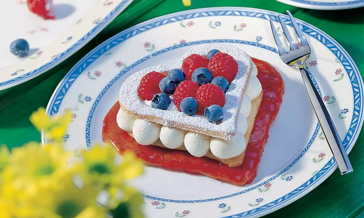 Ein herzförmiges Gebäck mit Puderzucker, Sahnetupfen und frischen Himbeeren sowie Heidelbeeren auf roter Fruchtsauce auf einem Teller