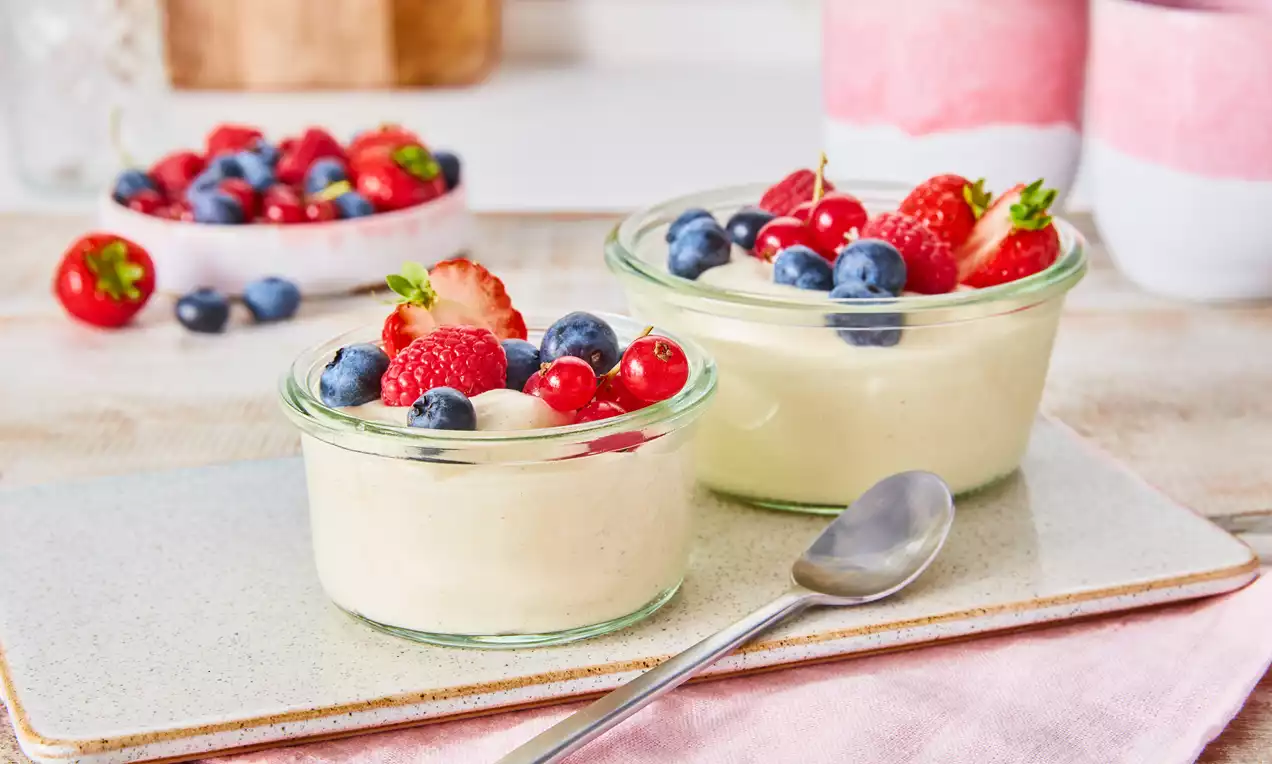 Vanille-Mousse in kleinen Gläsern, hellcremige Textur, dekoriert mit frischen roten und blauen Beeren auf hellem Teller mit silbernem Löffel