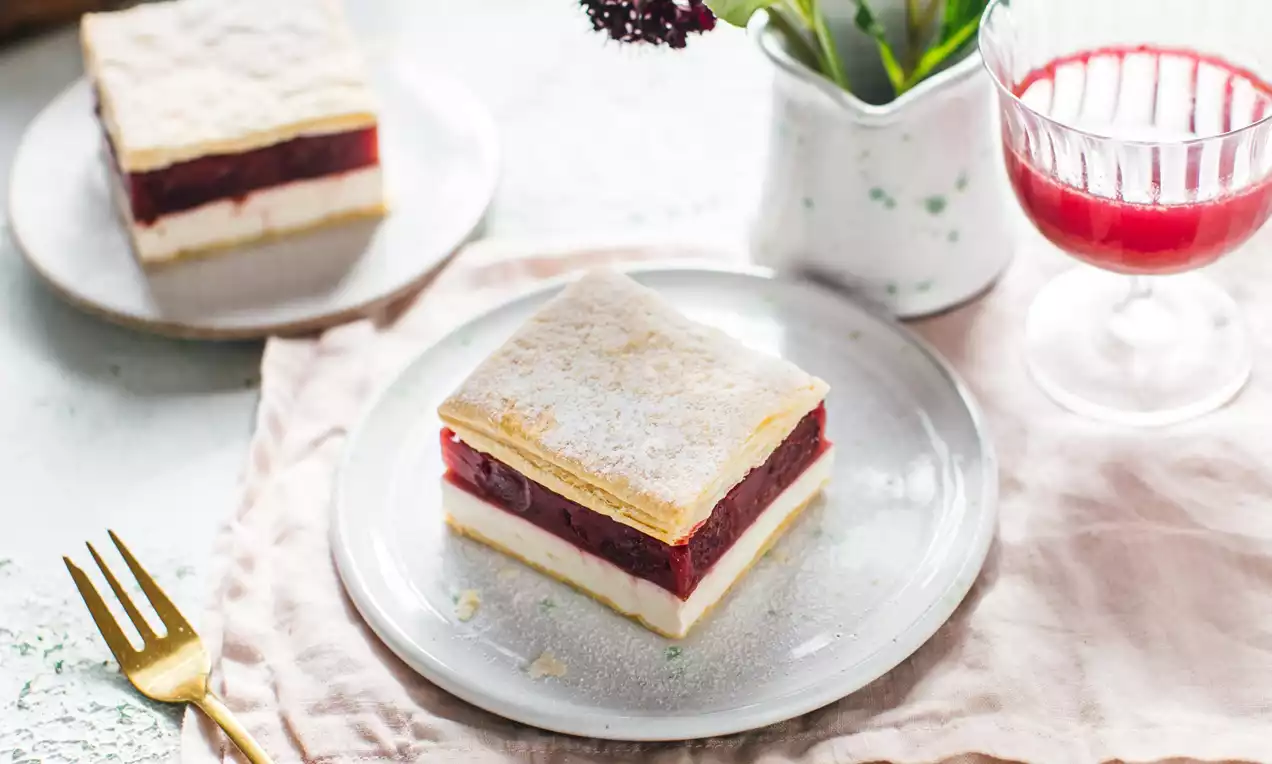 Tvarohovo-višňový zákusok s vrstvou bieleho tvarohu a lesklých červených višní medzi dvoma zlatistými lístkovými plátmi na bielom tanieri