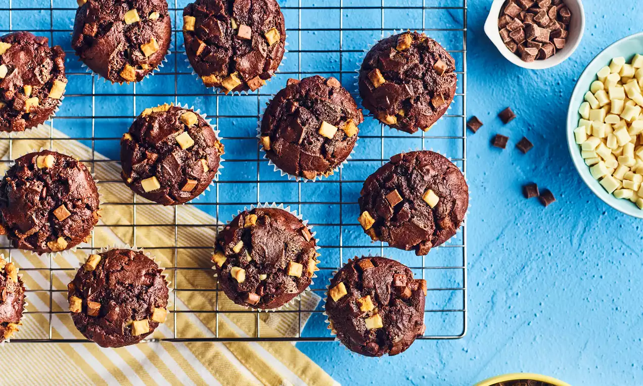 Triple chocolate muffins with rich brown tops, studded with dark, milk and white chocolate chunks, arranged on a wire rack over a blue surface