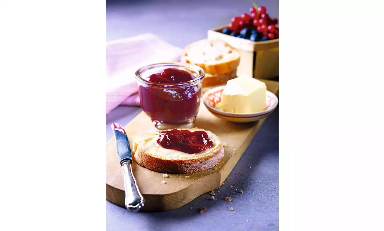 Zamatový džem tmavorubínovej farby v sklenenom pohári a na natretom chlebe, vedľa kus masla, noža a čerstvého ovocia na drevenej doske