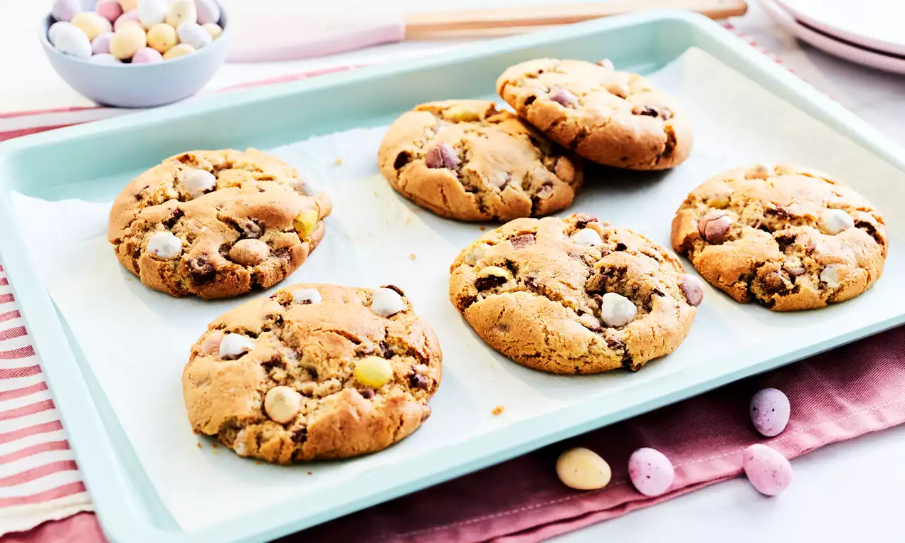 Golden brown cookies with a soft, textured surface speckled with pastel mini eggs, arranged on a pale turquoise tray with paper lining