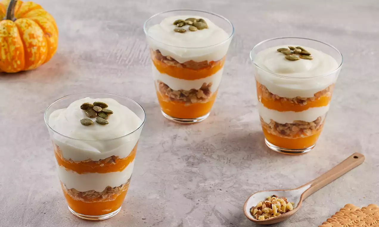 Kat kat düzenlenmiş bal kabaklı puding, beyaz krema ve çıtır granola ile doldurulmuş, üstü kabak çekirdekleriyle süslenmiş şeffaf bardaklar