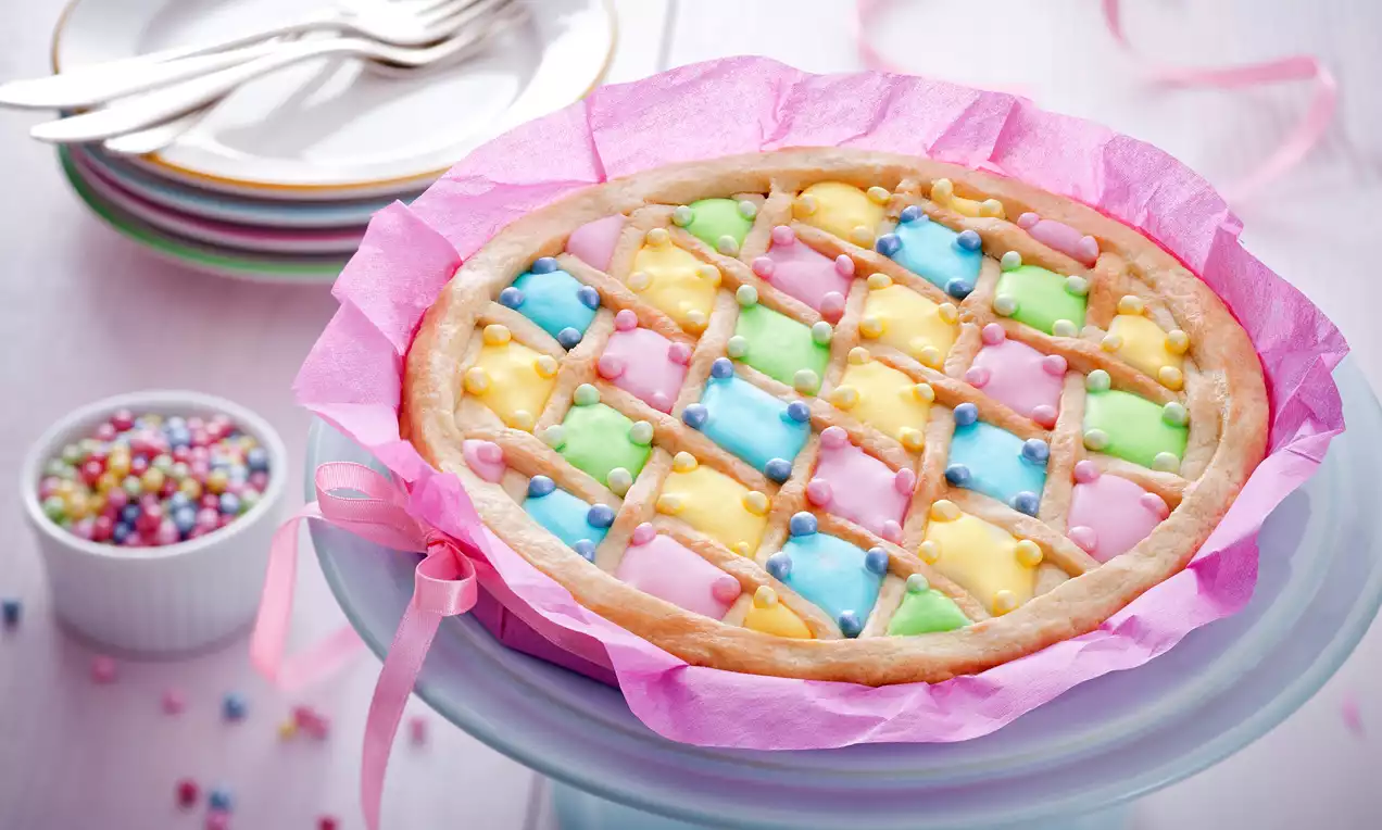 Mazurek na złocistym, kruchym spodzie w różowym papierze, udekorowany kratką z pastelowego lukru i białymi cukrowymi perełkami