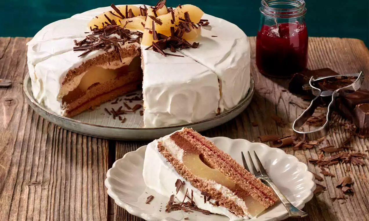 Eine Birnentorte mit weißer Sahneschicht, flaumigem Schokobiskuit und ganzer Birnenfüllung, dekoriert mit Schokoraspeln auf Holzuntergrund