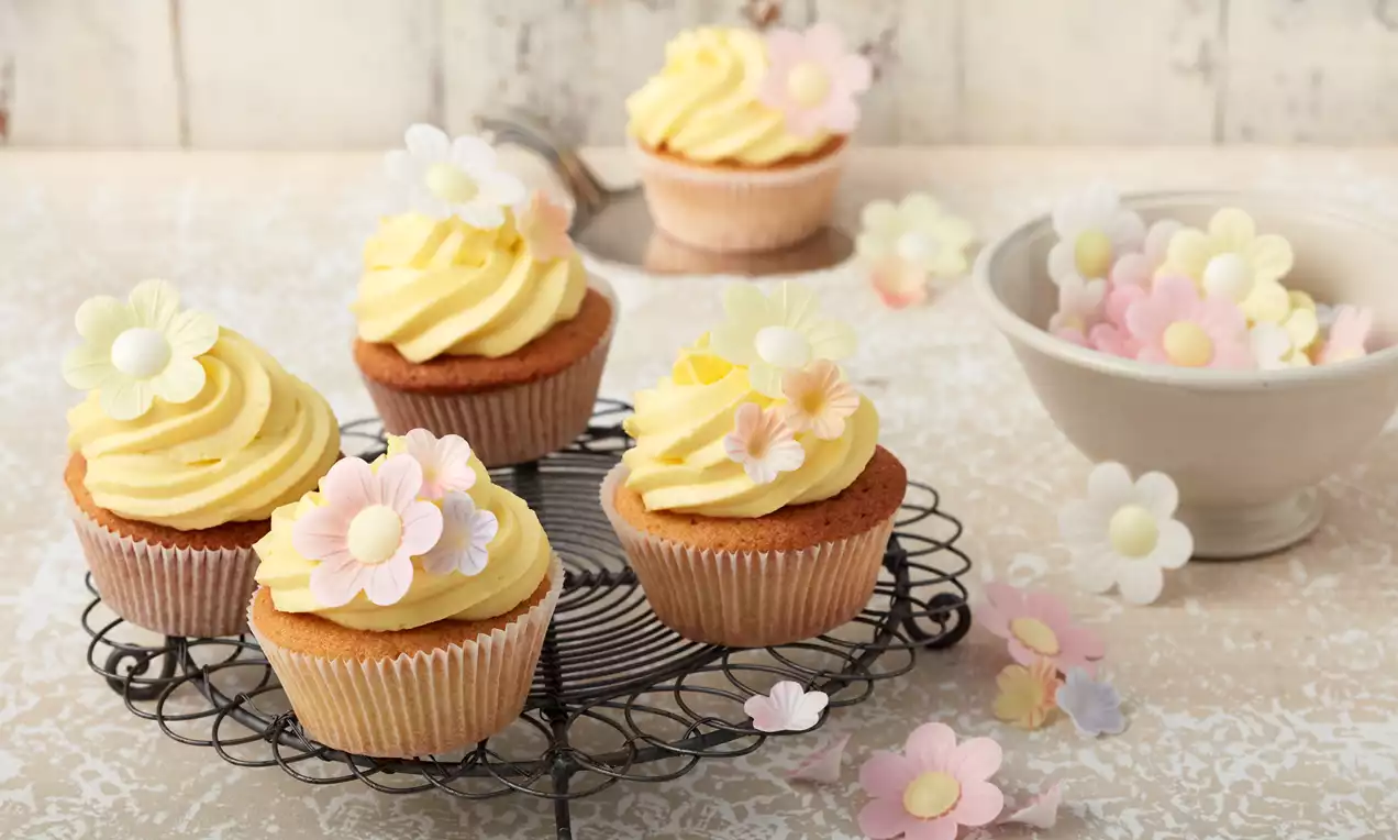 Zitronen-Cupcakes mit goldbraunem Teig und gelber Creme, verziert mit weißen und pastellfarbenen Blüten auf schwarzem Drahtgestell