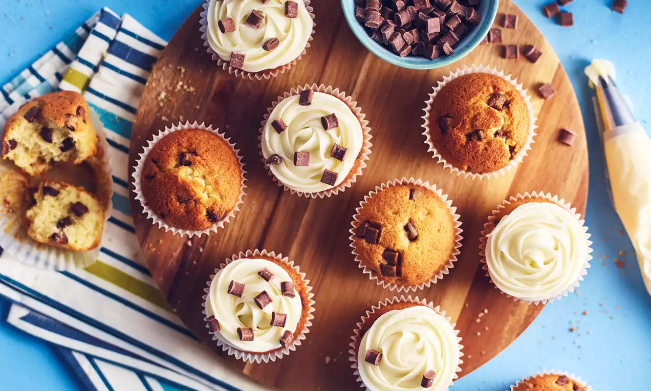 Golden fairy cakes topped with white swirls of cream and scattered chocolate chunks arranged on a round wooden plate with a bowl of chocolate pieces