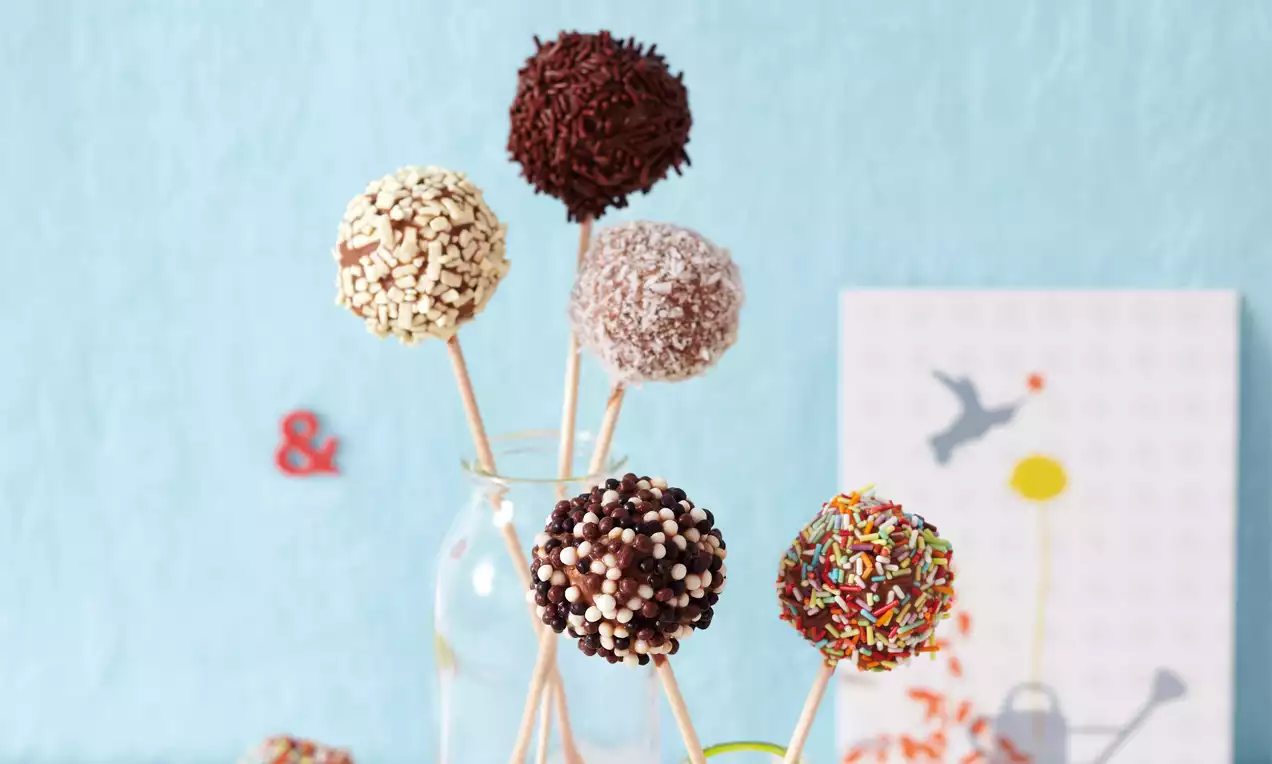 Barevné cake pops na dřevěných tyčinkách v skleněné váze, zdobené čokoládovými kuličkami, posypy a drobnými kousky, na světlém podkladu