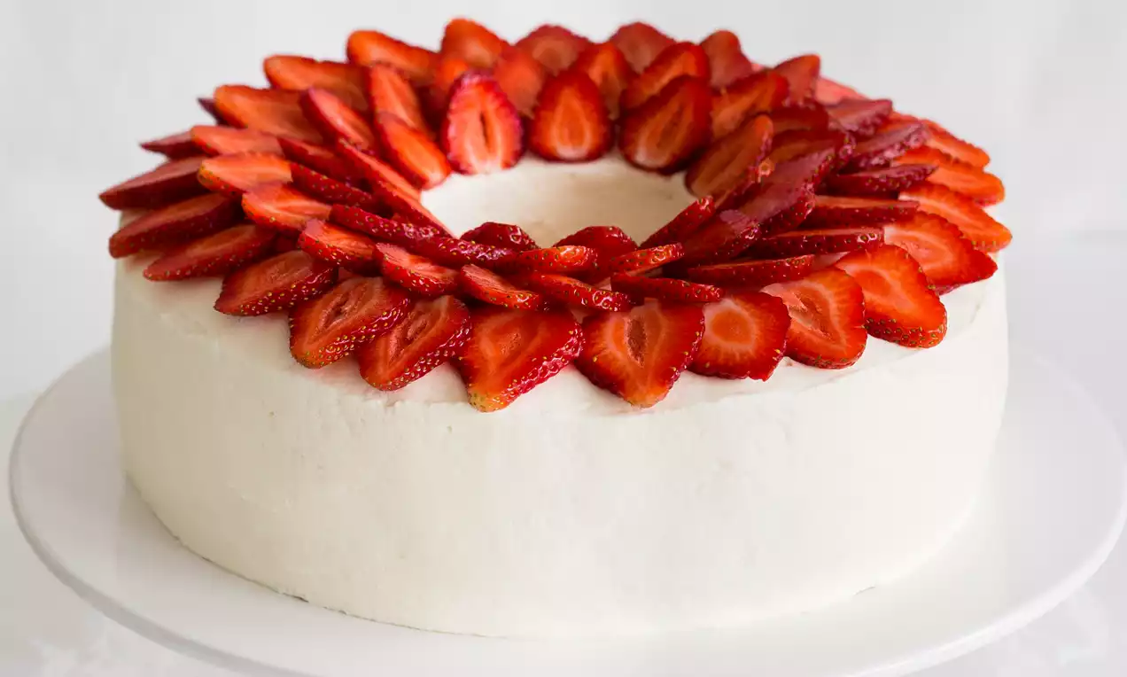 Biały tort w kształcie wianuszka udekorowany równomiernie ułożonymi plasterkami soczystych truskawek na białej paterze