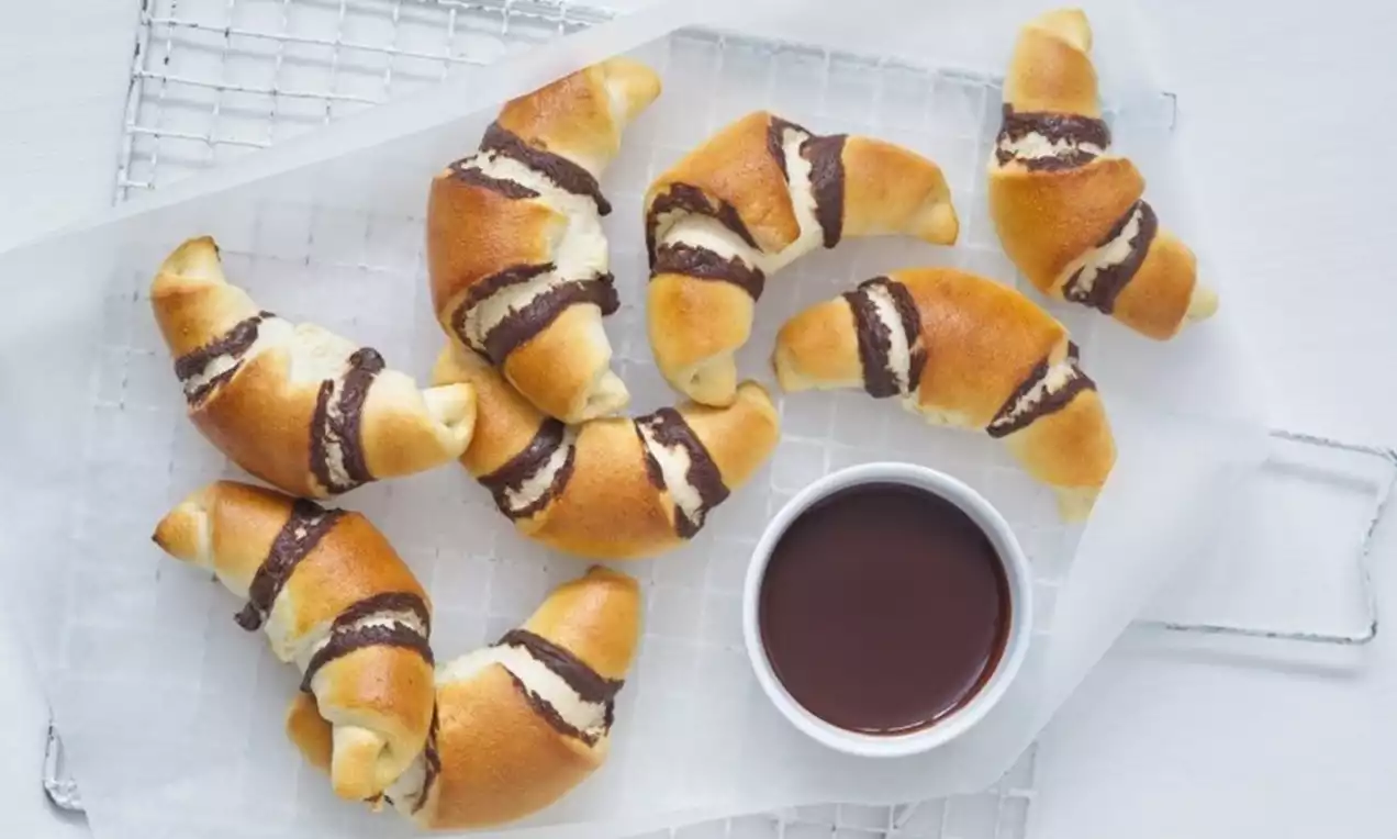 Zlatavé rohlíčky z listového těsta plněné tmavou čokoládou, položené na pečicím papíře s mřížkou a miskou čokoládové omáčky