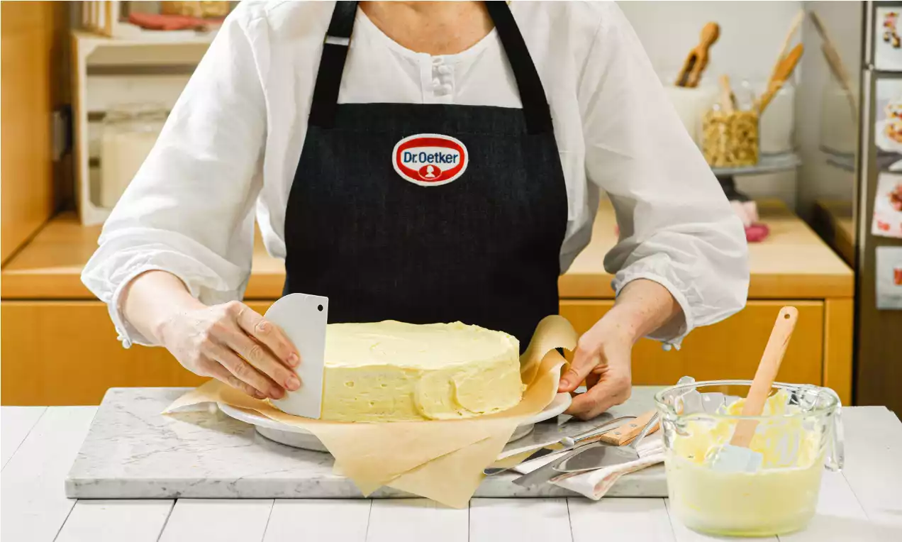 Eine Frau streicht eine hellgelbe Cremetorte mit einer Teigkarte ein, die Torte liegt auf Backpapier und einem weißen Teller auf Marmorplatte