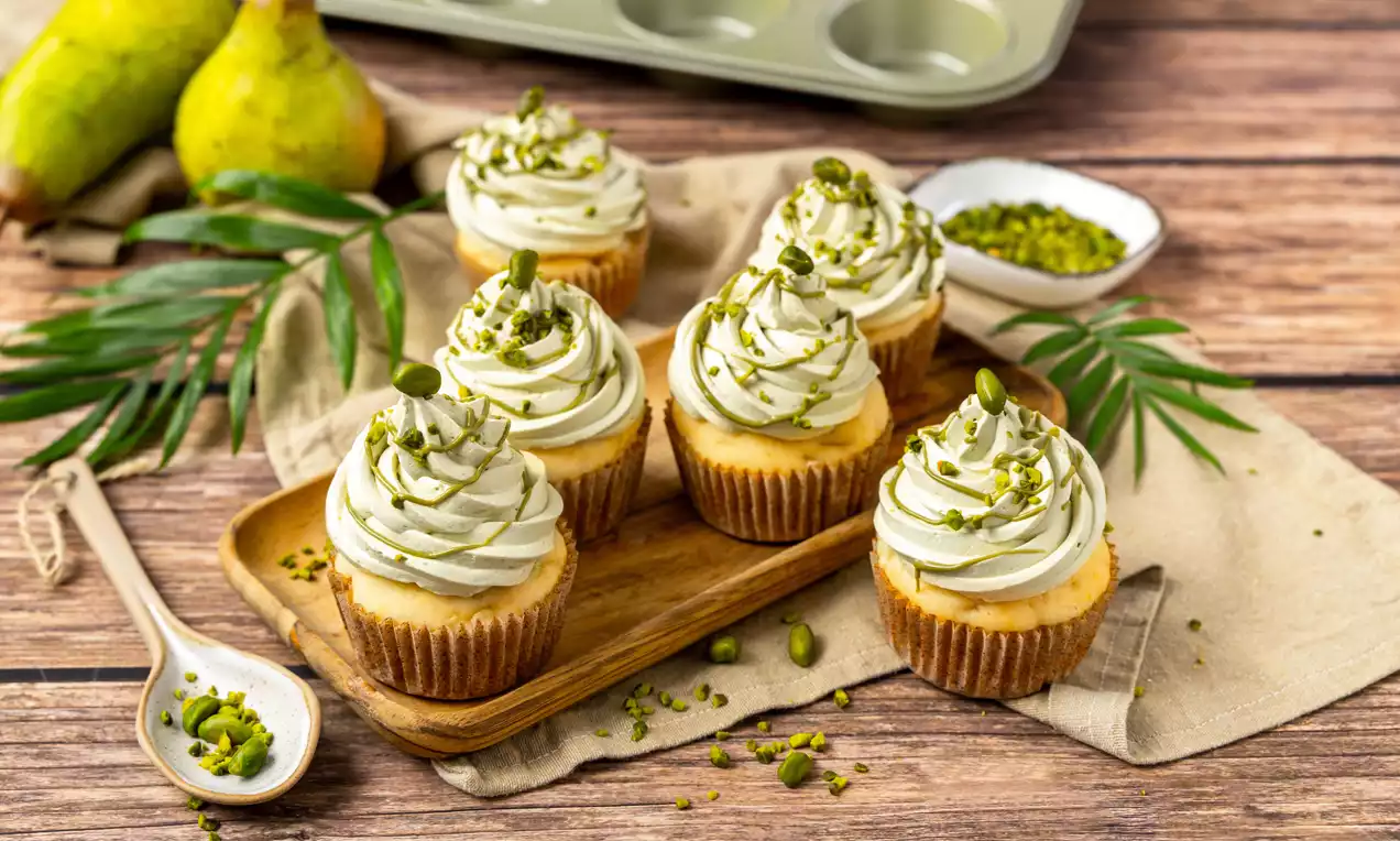 Vegane Birnen-Muffins mit hellem Rührteig, cremiger Pistazienhaube und gehackten Pistazien auf einem Holztablett, umgeben von Birnen und Blättern