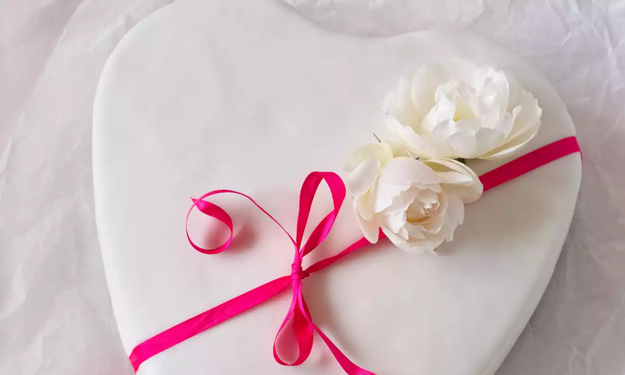Snehobiely tortový povrch v tvare srdca zdobený jemnými bielymi kvetmi a ružovou stuhou, položený na svetlom podklade