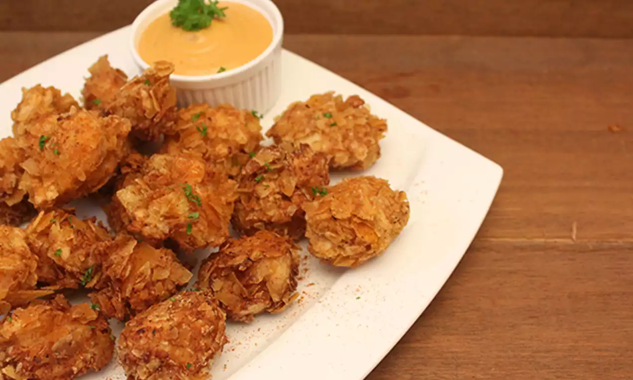 Golden-brown bite-sized chicken pieces with a crispy textured coating arranged on a white plate beside creamy dipping sauce in a ramekin