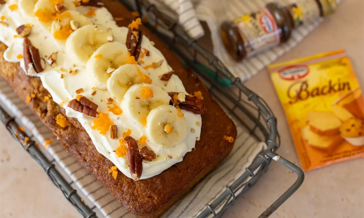 Langwerpige bruine cake met romige witte topping, plakjes banaan, oranje wortelrasp en pecannoten op een metalen rooster
