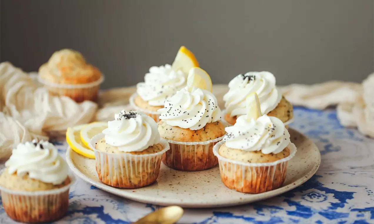 Citronovo-makové cupcakes zdobené bílým krémem, posypané mákem, s plátky citronu na keramickém talíři na modrobílém ubrusu
