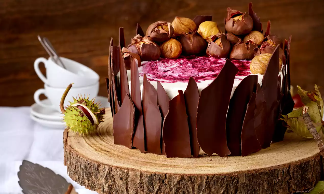 Runde Torte mit weißer Creme, violett marmoriertem Fruchtspiegel, umhüllt von dunkler Schokolade und dekoriert mit glänzenden Maroni, auf Holzplatte angerichtet