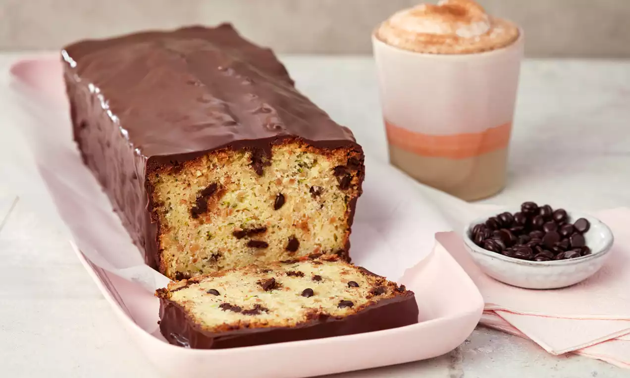 Länglicher Zucchinikuchen mit glänzender Schokoglasur, angeschnitten auf einer rosa Platte, daneben Cappuccino und Schüssel mit Kaffeebohnen