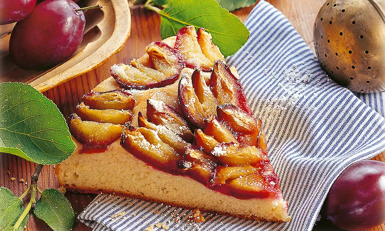 Fetta di torta dorata con superficie di prugne rosso-scuro adagiate a ventaglio, spolverata di zucchero a velo su tessuto a righe bianche e blu