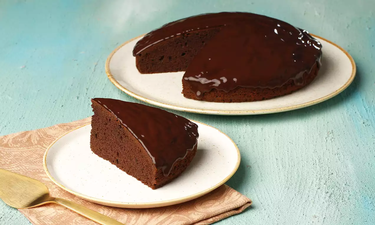 Pürüzsüz çikolata sosuyla kaplı yuvarlak kek, bir dilimi sade beyaz tabakta açık mavi zeminde ve altın renkli çatal eşliğinde