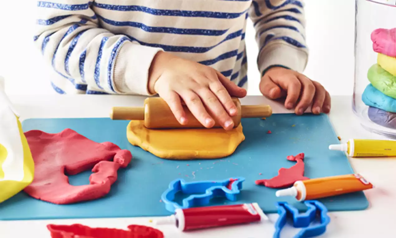 Child's hands rolling yellow playdough with a wooden rolling pin on a blue mat surrounded by colorful playdough cutters and tubes
