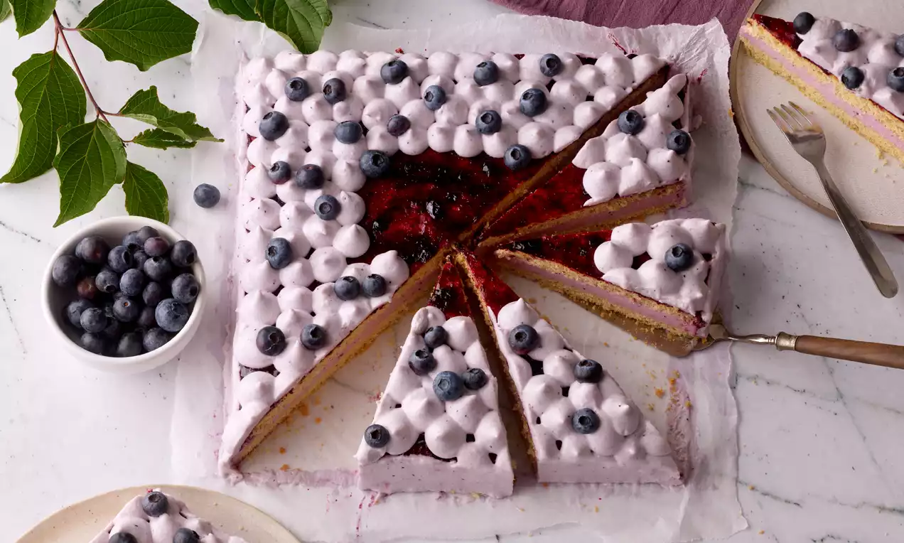 Quadratischer Kuchen mit violetter Creme, frischen Blaubeeren und roten Fruchteinlagen, in Stücke geschnitten, auf weißem Papier präsentiert