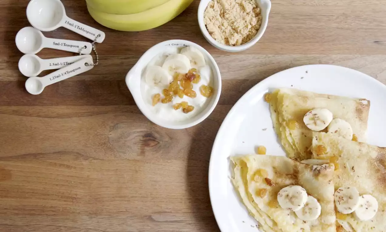 Goudgele pannenkoeken gevouwen op een wit bord met plakjes banaan en rozijnen, naast een schaaltje plattekaas en een tros bananen