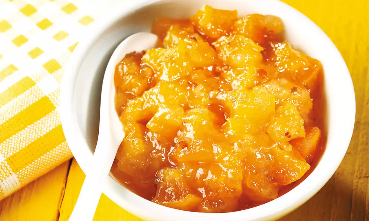 Hrubo nasekané kúsky hrušiek a marhúľ v sýto oranžovom lesklom sirupe, podávané v bielej miske s lyžicou na žltom podklade