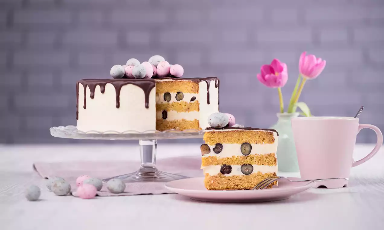 Drip-Cake mit weißer Creme, dunklem Schokoladenguss, frischen Heidelbeeren und pastellfarbenen Deko-Elementen auf einer Etagere