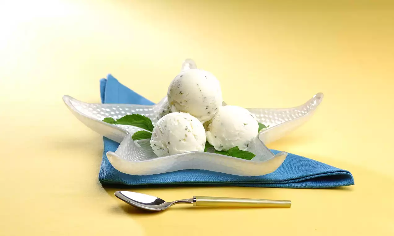 Parlak cam tabakta üç top keskin yeşil naneyle süslenmiş beyaz dondurma, mavi bir bez ve altın saplı kaşıkla sunuluyor