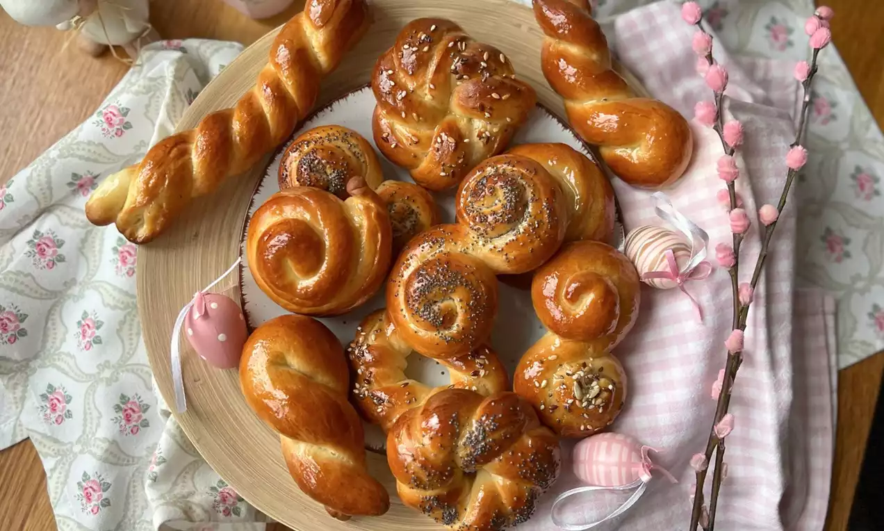 Lesklé zlatohnědé jidáše různých zakřivených tvarů, posypané mákem a semínky, na světlém dřevěném talíři s velikonočními dekoracemi a větvičkami