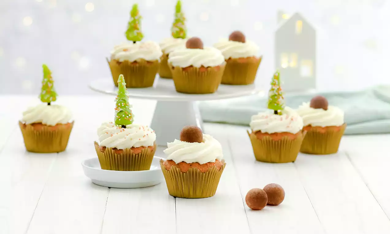 Zlatnosmeđi muffini u papirnatim kalupima s kremastim bijelim preljevom, ukrašeni zelenim božićnim drvcem i čokoladnom kuglicom