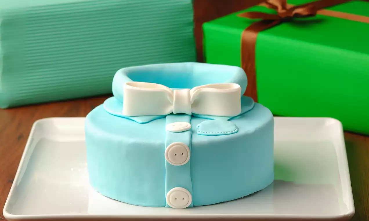 Açık mavi gömlek şeklinde tasarlanmış, beyaz düğmeler ve fiyonk detaylarıyla süslenmiş pasta, beyaz dikdörtgen tabakta sunulmuş