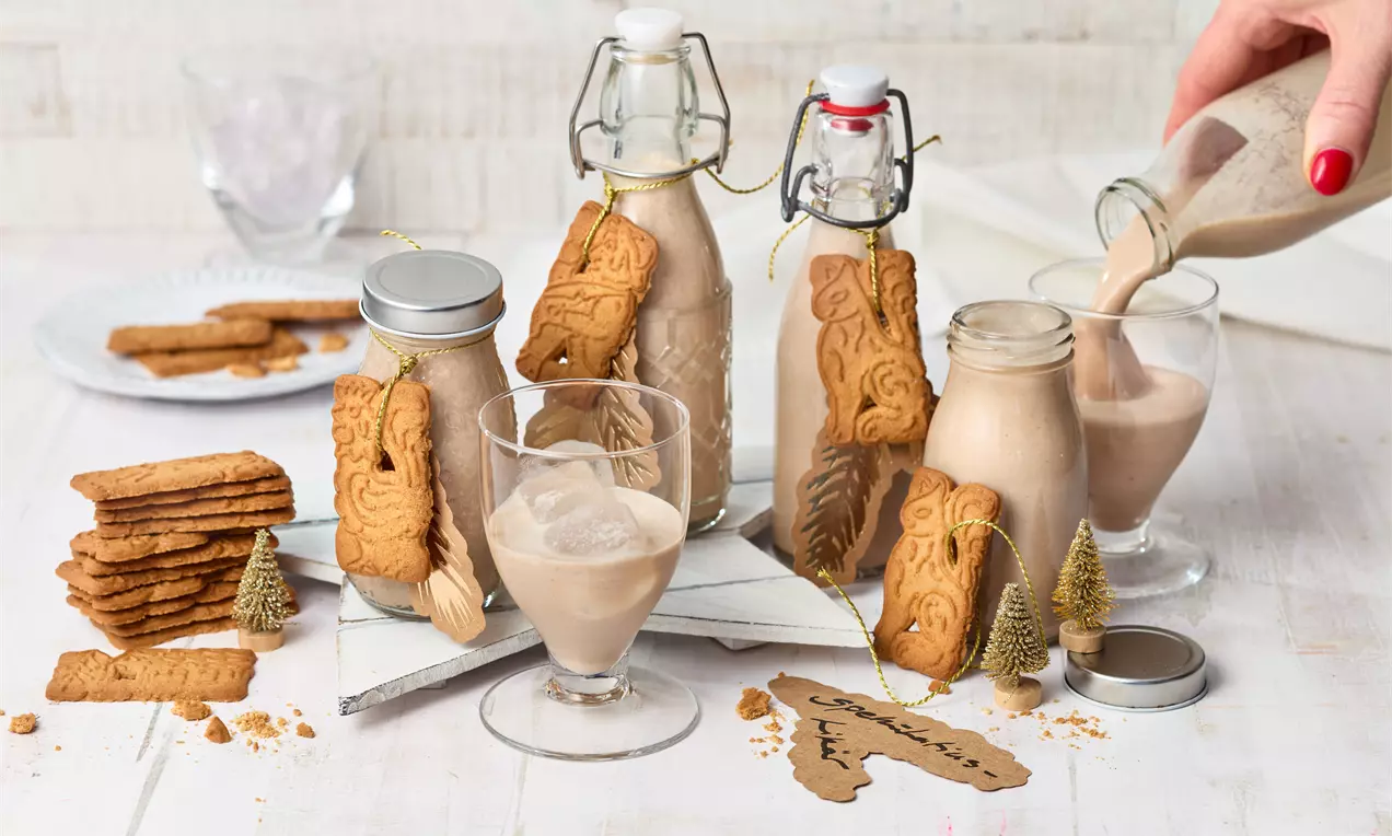 Spekulatiuslikör in Glasflasche mit goldener Kappe, cremig hellbraun, dekoriert mit Spekulatius-Keksen, Zimtstange und Tannengrün