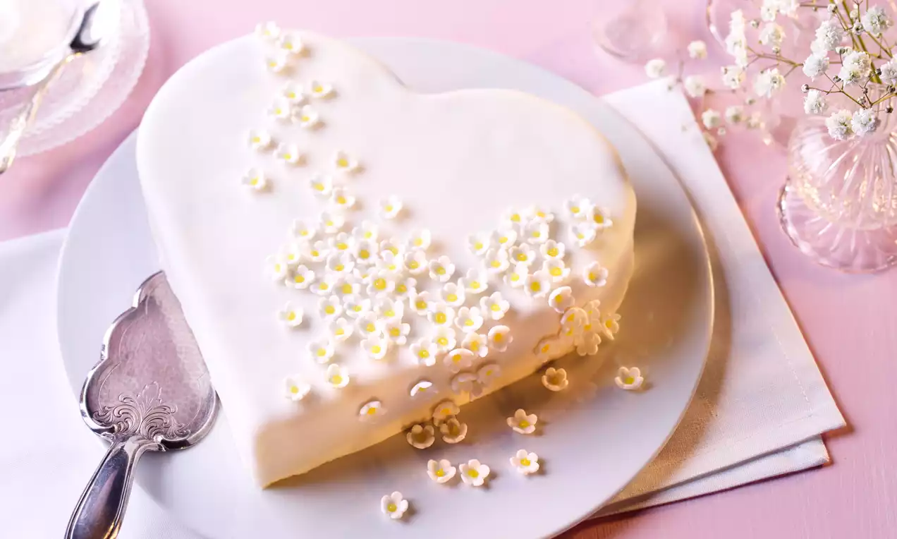 Herzförmige Torte mit glatter weißer Glasur, dekoriert mit kleinen weißen Blüten und gelben Mitte auf einer weißen Platte