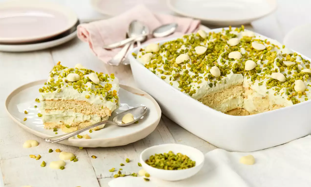 Ein Stück Pistazien-Tiramisu mit heller Creme und grünen Pistazienstücken auf einem beigefarbenen Teller neben einer weißen Auflaufform