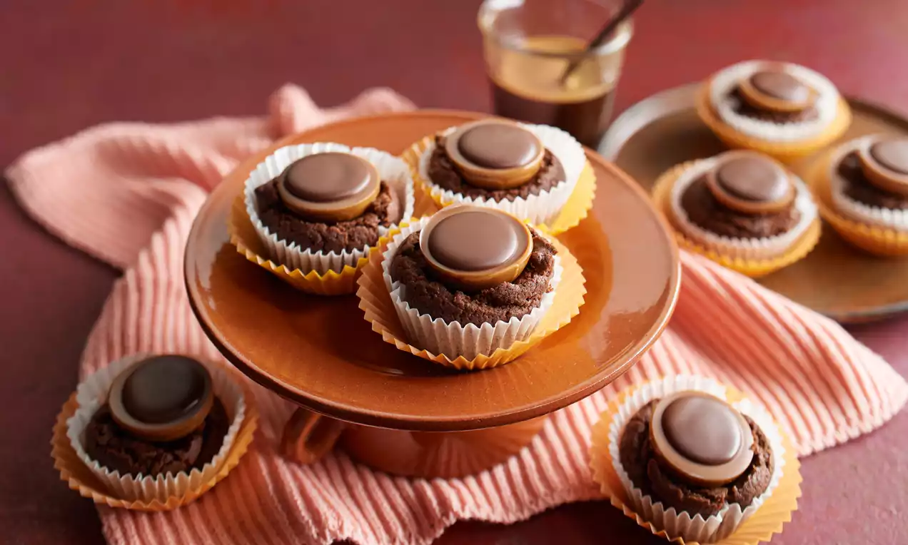 Saftige Brownies mit glatter Toffifee®-Creme, dunkelbraun und rundlich, in gelben Papierförmchen auf brauner Tortenplatte angerichtet