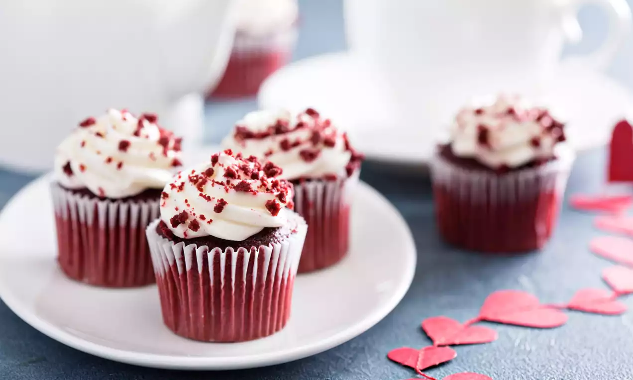 Four vibrant red velvet cupcakes with creamy swirls of white frosting and red crumbs on a white plate, surrounded by red paper hearts